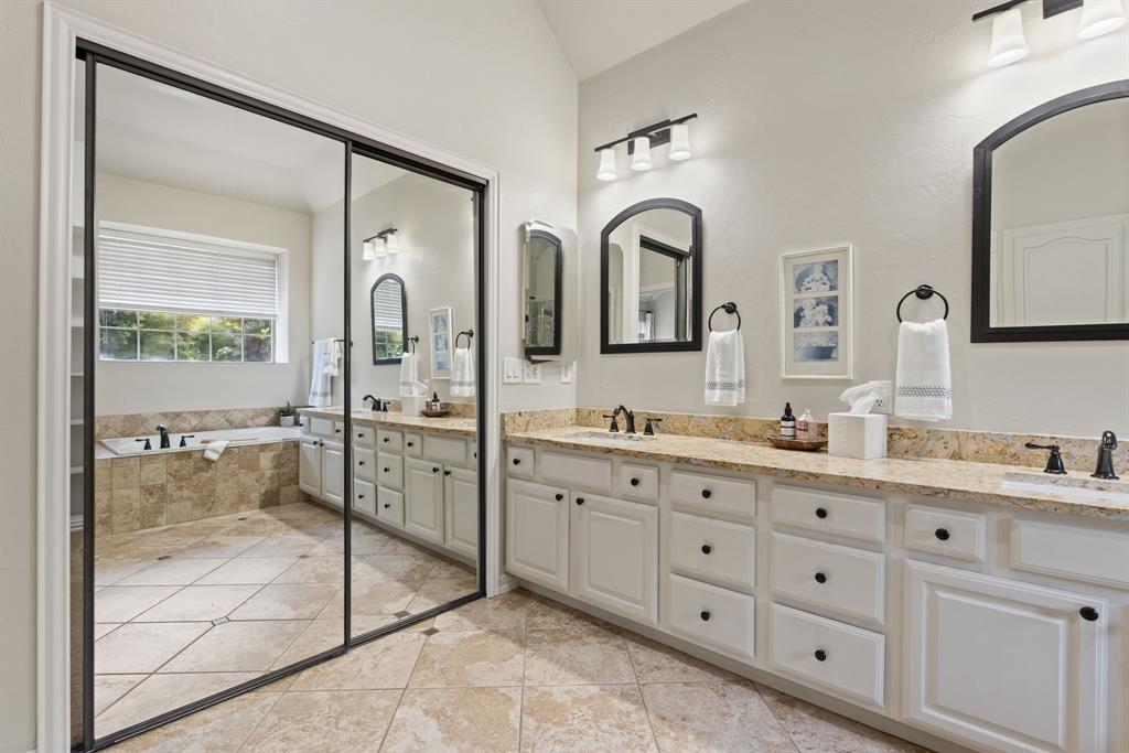 Dual vanity with stone-finish countertop, white shaker-style cabinetry, arched wall mirrors, stone tile flooring, and a jetted soaking tub with tile surround