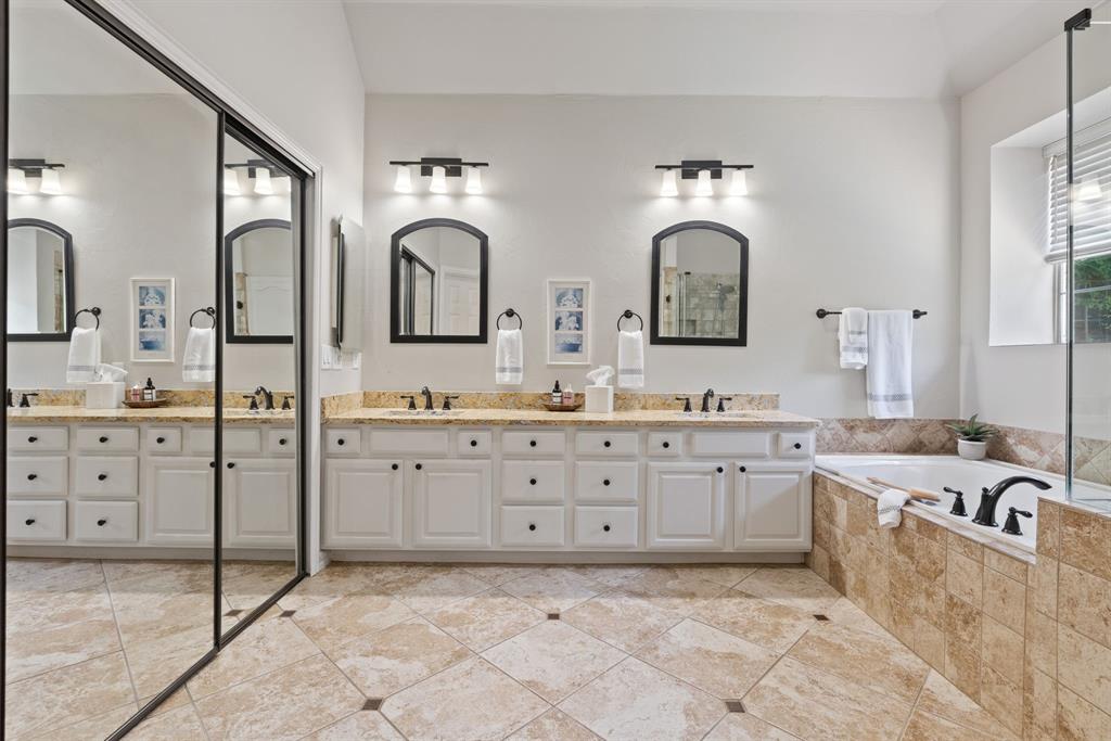 Dual-sink vanity with stone countertop, large-scale mirror wall, tile-surround soaking tub, and tile flooring with decorative inlay