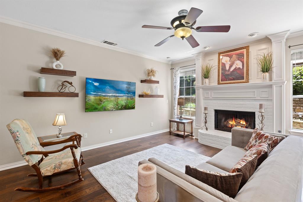 Living area featuring dark wood-finish flooring, a white brick fireplace with a custom mantel, built-in floating shelves, crown molding, and a ceiling fan