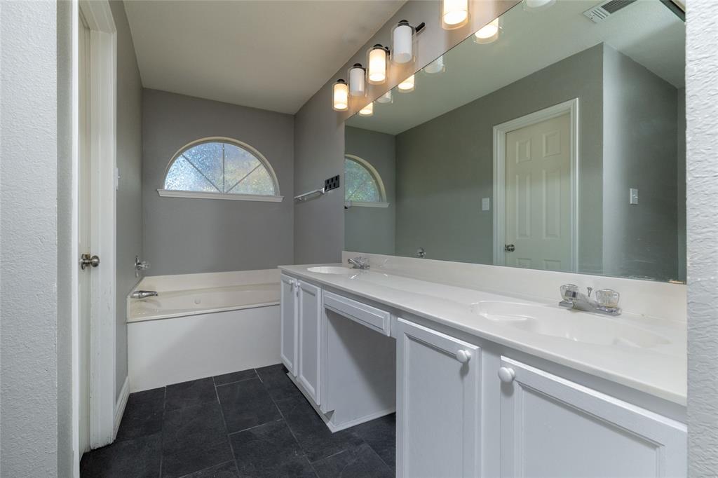 Dual-sink vanity with white cabinetry and a solid-surface countertop, full-width wall mirror, soaking tub, arched window, and stone-finish tile flooring