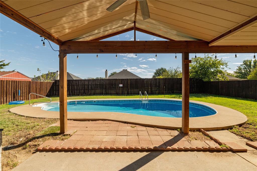 Covered patio with wood-beam construction, ceiling fan, and string lighting, featuring a backyard pool with concrete surround and full wood fencing