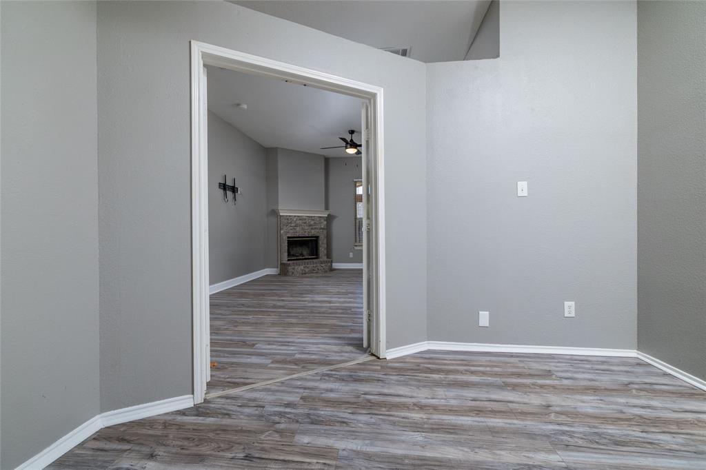 Interior room featuring wood-finish flooring, neutral wall tones, and a doorway leading to a space with a floor-to-ceiling stone fireplace and a ceiling fan Interior room featuring wood-finish flooring, neutral wall tones, and a doorway leading to a space with a floor-to-ceiling stone fireplace and a ceiling fan