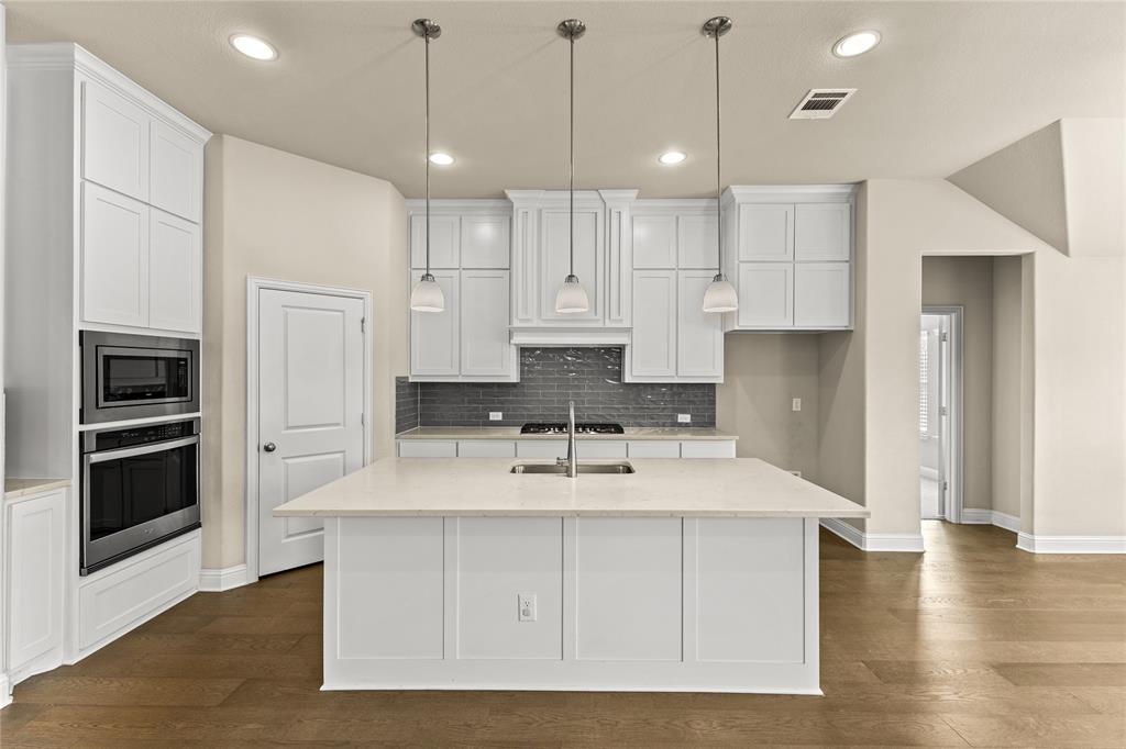 Kitchen featuring a large central island with light-toned solid surface countertops, white shaker-style cabinetry, a dark tiled backsplash, stainless steel built-in double ovens, and three pendant light fixtures over the island