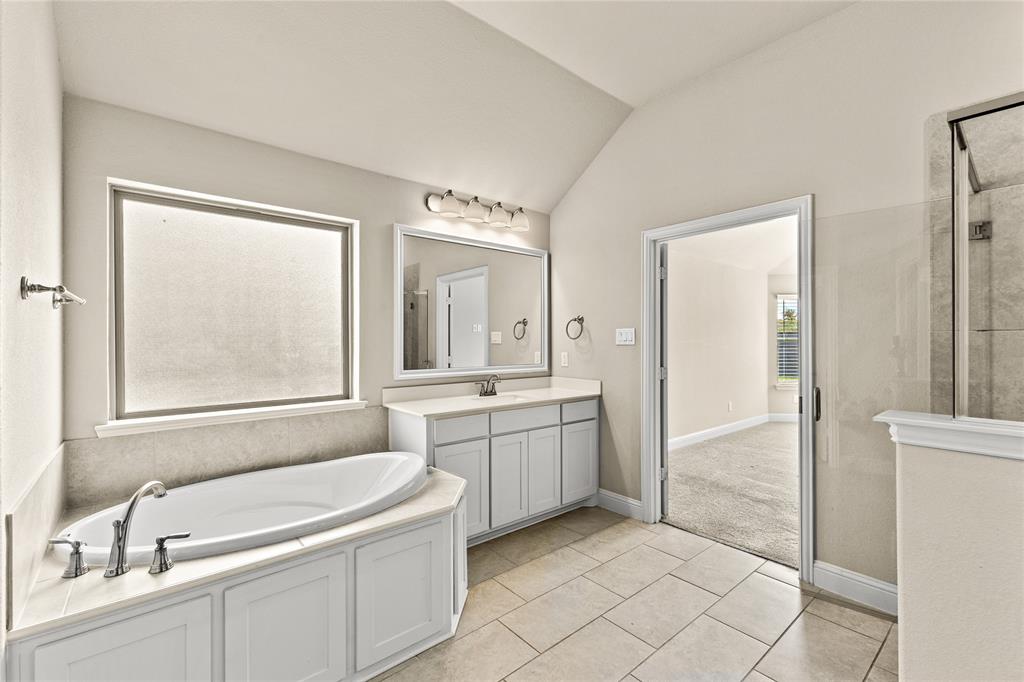 Built-in soaking tub with a tiled surround, dual-sink vanity with white cabinetry, framed vanity mirror, glass-enclosed shower, and neutral tile flooring