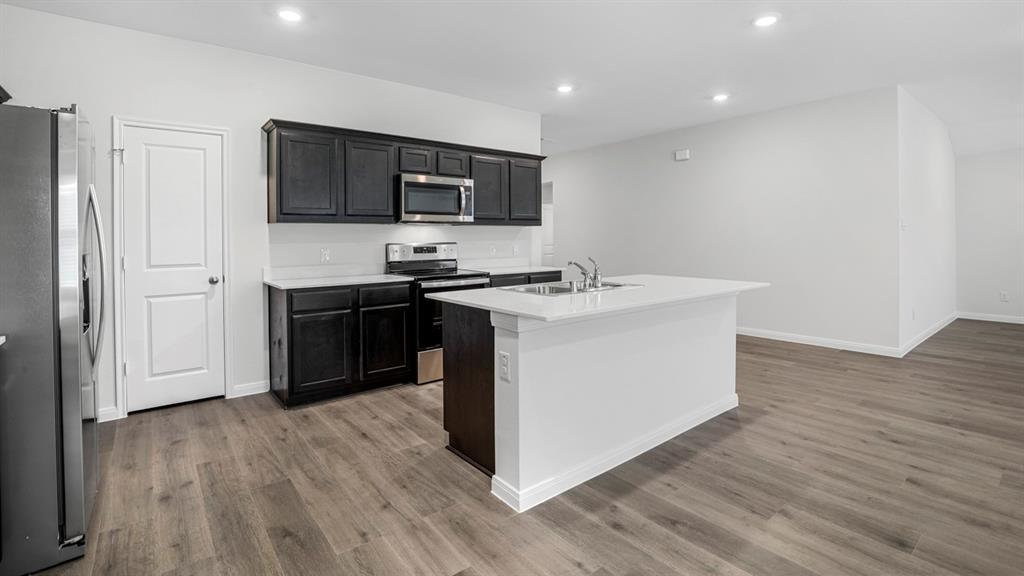 Modern kitchen featuring dark wood cabinetry, stainless steel appliances, a white island with an integrated sink, light countertops, and wood-finish flooring