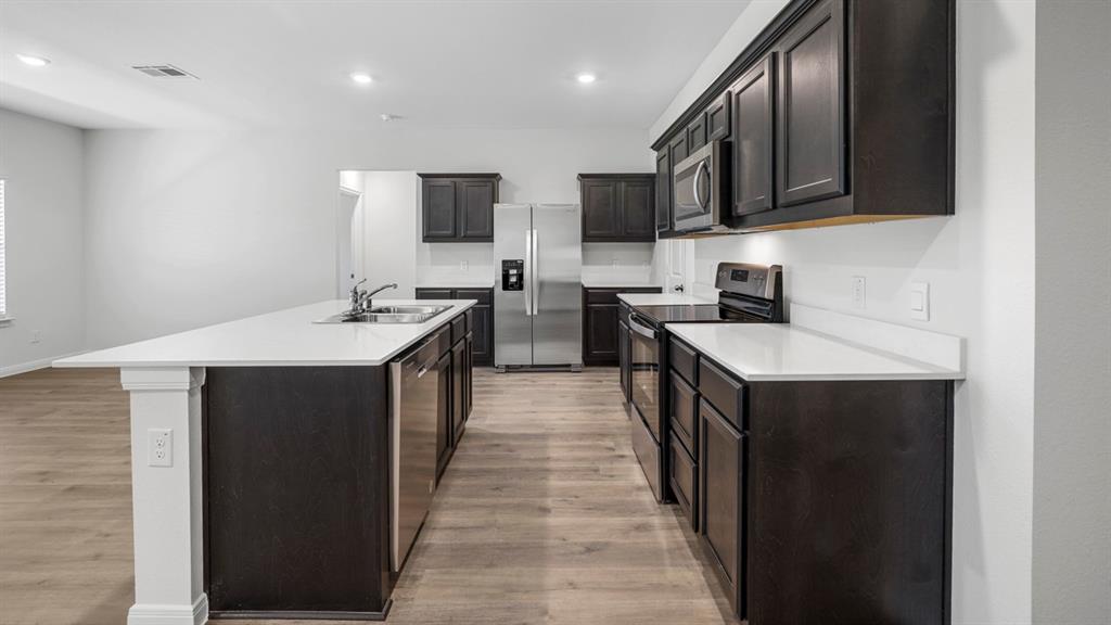 Kitchen featuring an elongated island with a double basin sink, dark cabinetry, light-toned countertops, stainless steel appliances, and wood-finish flooring