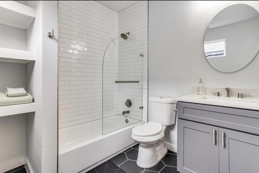 Bathroom featuring a gray vanity with a stone countertop, white subway tile shower surround with an arched glass panel, built-in shelving, and black hexagonal floor tile