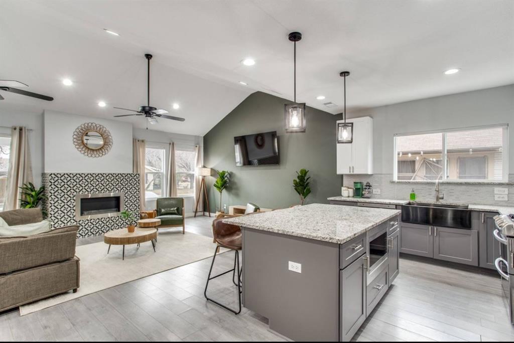 Open-concept kitchen and living area featuring vaulted ceilings, wood-finish flooring, a patterned tile fireplace surround, an expansive center island with stone countertop, and charcoal-toned cabinetry Open-concept kitchen and living area featuring vaulted ceilings, wood-finish flooring, a patterned tile fireplace surround, an expansive center island with stone countertop, and charcoal-toned cabinetry