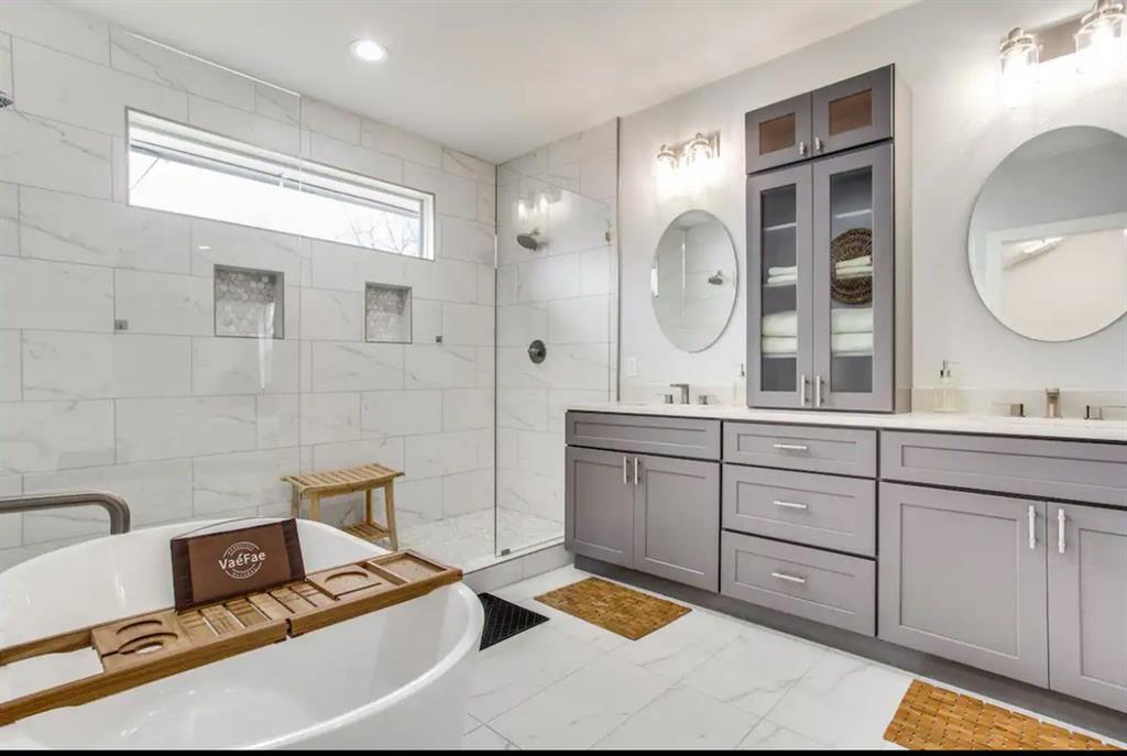 Freestanding soaking tub, double-sink vanity with gray shaker-style cabinetry, glass-enclosed shower with tiled surround, built-in linen tower, and recessed shower niches