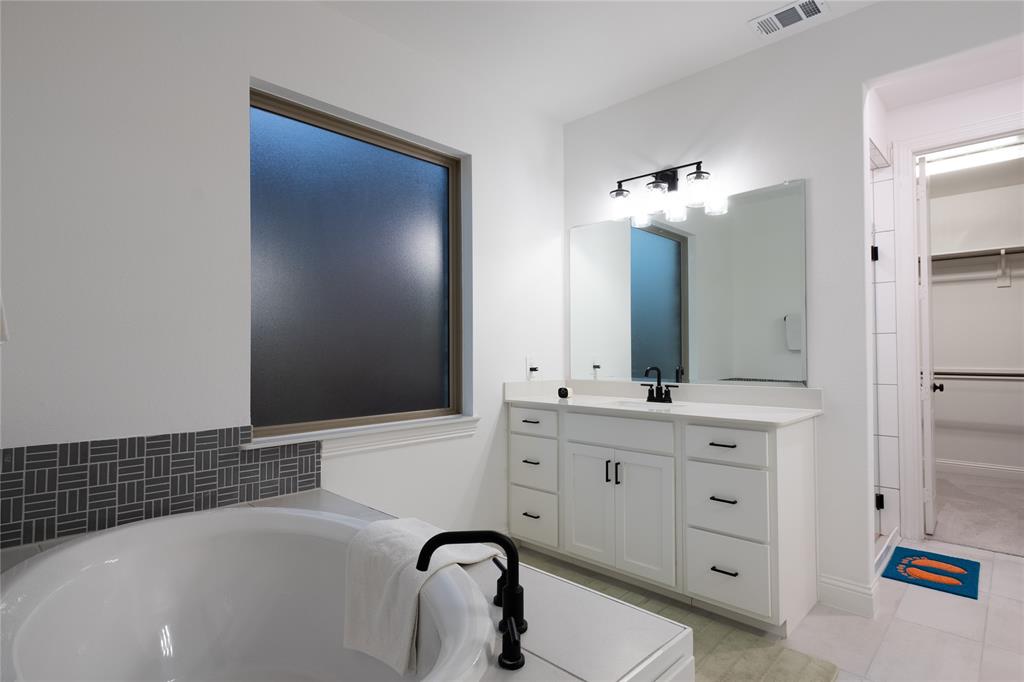 Built-in white vanity with stone-style countertop, undermount sink with matte black hardware, frosted window, deep soaking tub with matte black faucet, and patterned decorative tile backsplash