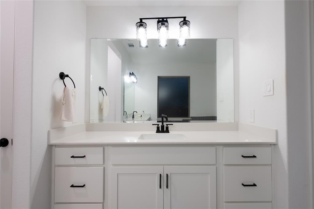 Bathroom vanity featuring white cabinetry, a solid-surface countertop with an integrated basin, a large plate glass mirror, matte black hardware, and a three-light fixture with textured glass shades