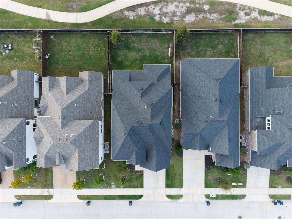 Aerial perspective featuring a residential lot with a paved driveway, concrete walkway, private fenced backyard, and shingled roofline