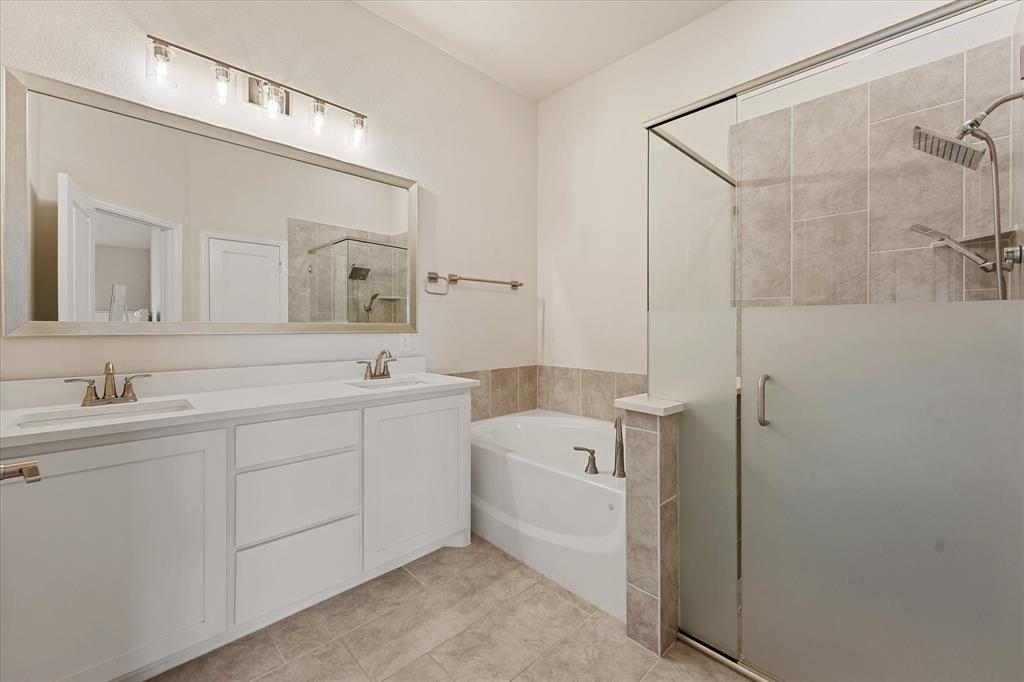 Bathroom featuring a dual-sink vanity with white cabinetry, a full-width vanity mirror, a separate soaking tub, and a walk-in shower with tile surround and frosted glass door