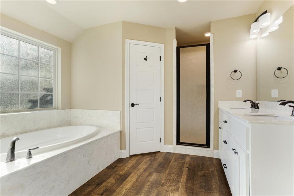 Bathroom featuring a deep soaking tub with a stone-finish surround, dual-sink vanity with light-toned cabinetry, wood-finish flooring, glass-enclosed shower, and multi-light wall sconce