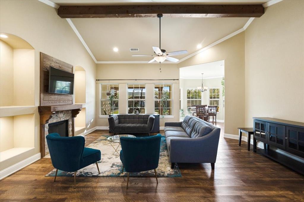 Living room featuring vaulted ceilings, a central exposed wood beam, wood-finish flooring, a stone-surround fireplace with a wooden mantel, and built-in recessed shelving