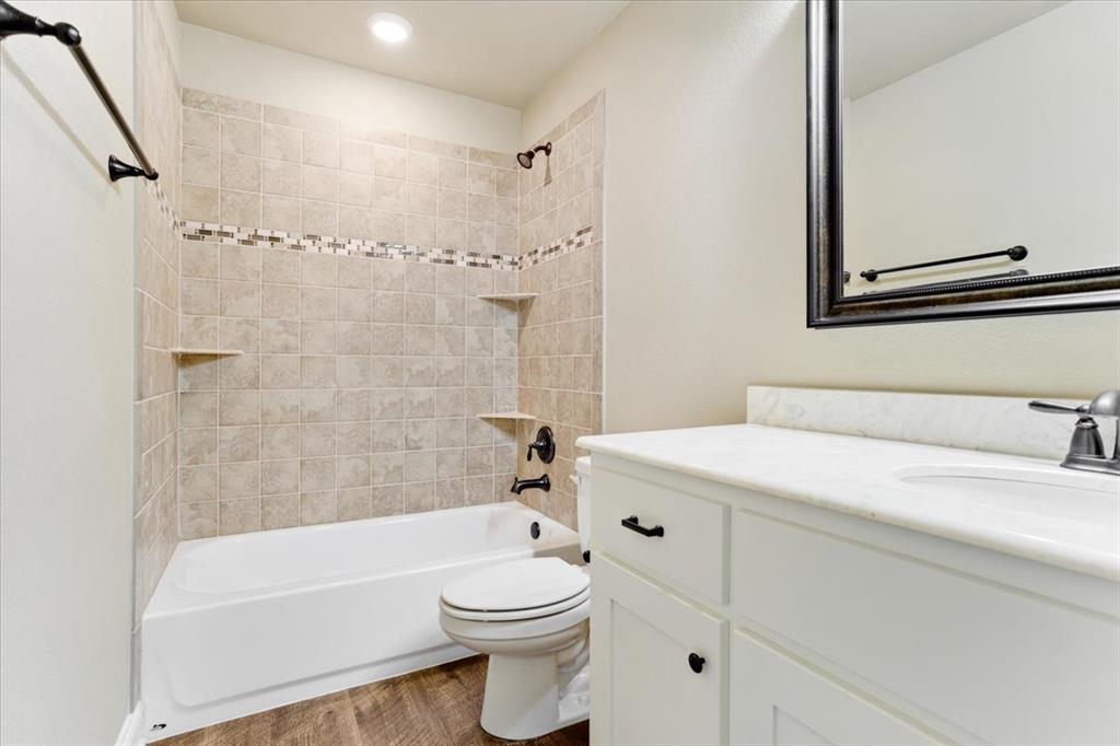 White vanity with stone-finish countertop, undermount sink, oil-rubbed bronze hardware, tub-shower combination with decorative tile inlay, and wood-finish flooring