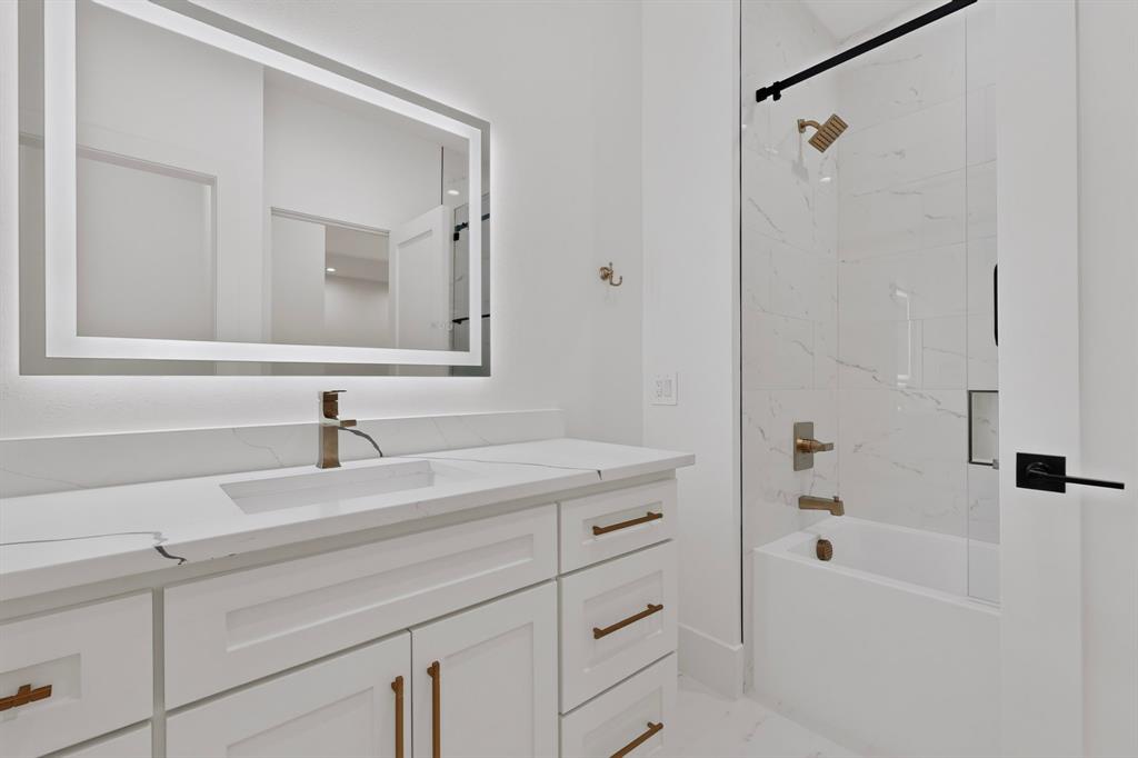 Shaker-style white vanity with a quartz countertop, integrated LED-backlit mirror, brass-finish hardware, and a tub-shower combination featuring tile surround and a frameless glass door