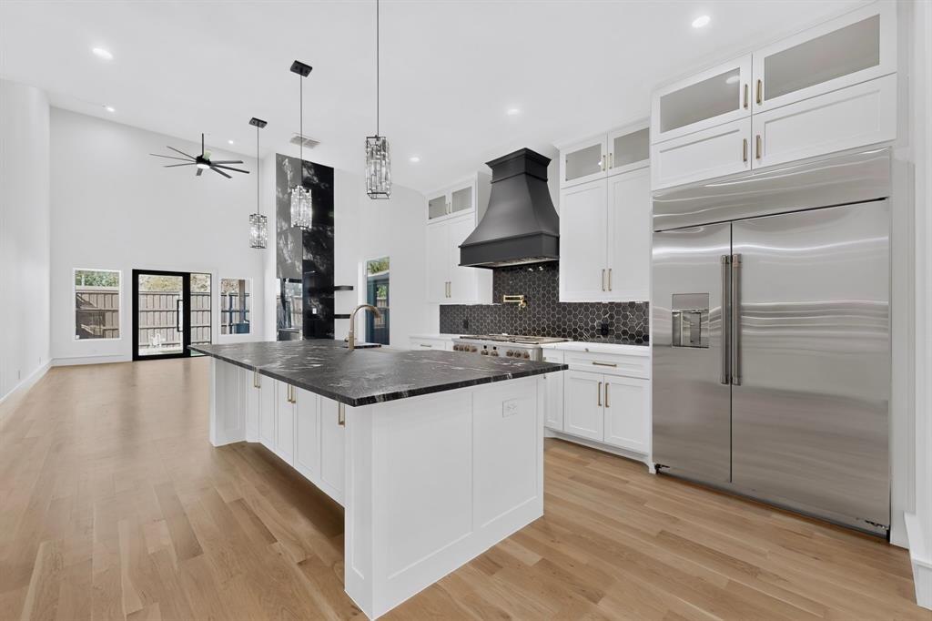 Kitchen featuring a center island with a dark stone countertop, white cabinetry with stacked uppers, a black range hood, stainless steel refrigerator, and wood-finish flooring