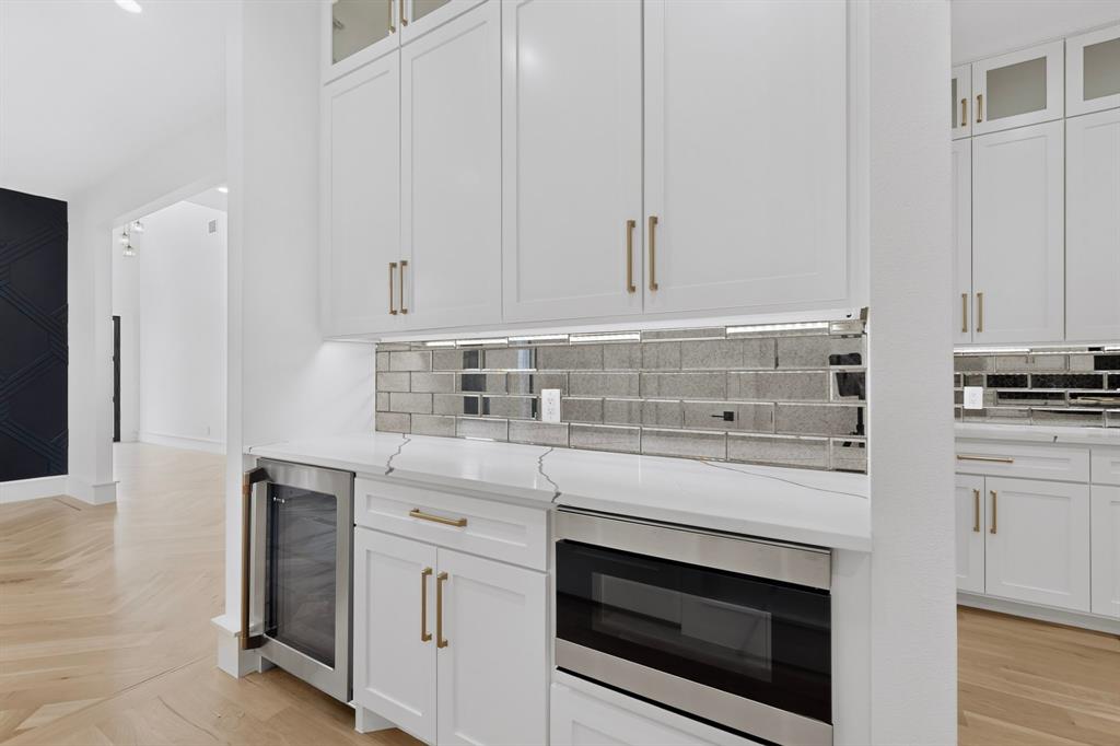 Shaker-style cabinetry with gold-toned hardware, stone-surface countertop, mirrored subway-tile backsplash, stainless steel under-counter beverage refrigerator, and drawer-style microwave