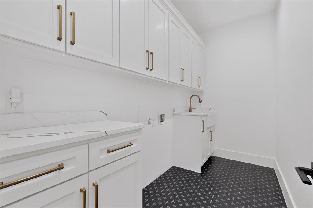 Laundry room featuring white shaker-style cabinetry, stone-finish countertops, apron-front utility sink, gold-tone hardware, and patterned tile flooring