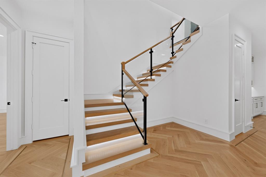 Open-riser staircase featuring wood-finish treads with integrated lighting, glass panels, a wood handrail, and black metal supports, complemented by herringbone-patterned wood-finish flooring Open-riser staircase featuring wood-finish treads with integrated lighting, glass panels, a wood handrail, and black metal supports, complemented by herringbone-patterned wood-finish flooring