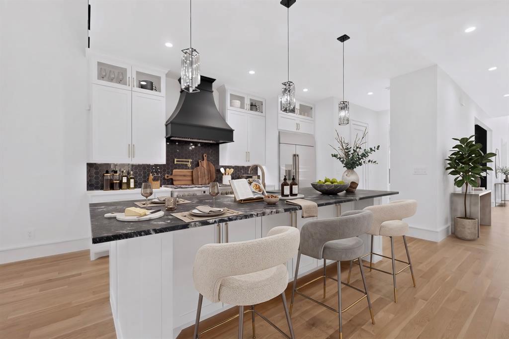 Kitchen featuring white shaker-style cabinetry, dark stone-finish island countertop, matte black statement range hood, geometric tile backsplash, and wood-finish flooring Kitchen featuring white shaker-style cabinetry, dark stone-finish island countertop, matte black statement range hood, geometric tile backsplash, and wood-finish flooring