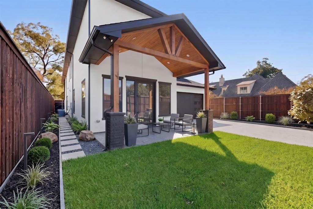 Covered patio featuring a wood-plank ceiling and recessed lighting, grounded by a concrete base and flanked by dark masonry pillars, complemented by a landscaped perimeter of stone pavers and lawn