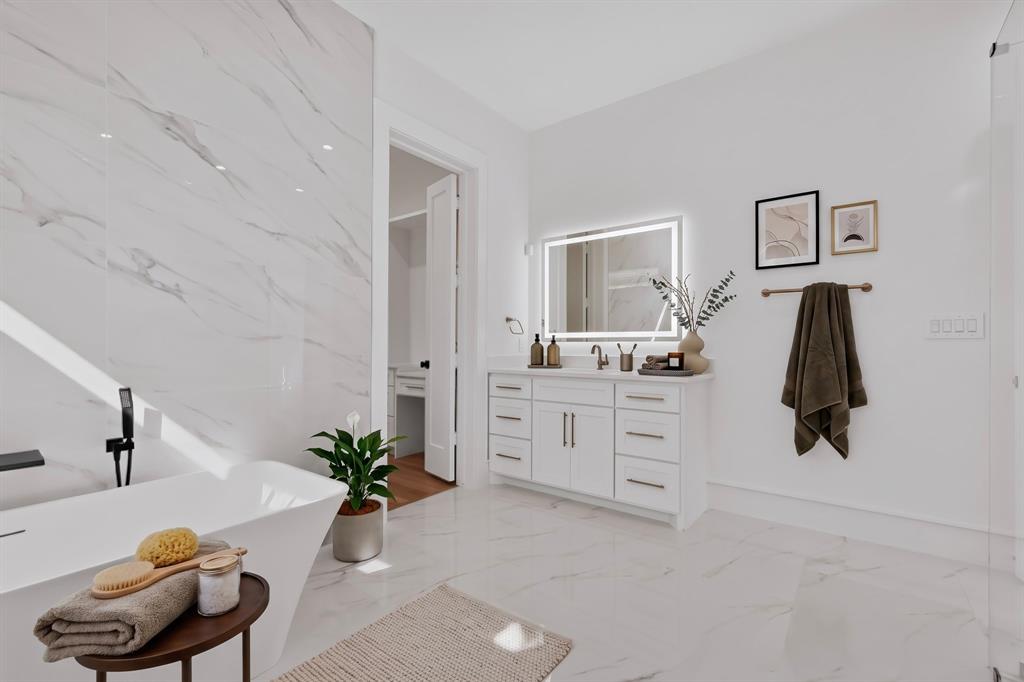 Freestanding soaking tub with a wall-mounted faucet, large-format stone-finish tile flooring, white shaker-style vanity with metal hardware, and an illuminated wall-mounted mirror