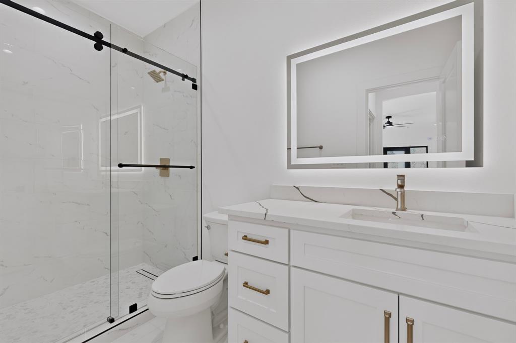 Walk-in shower with floor-to-ceiling veined tile, frameless glass sliding enclosure, brass-finish fixtures, quartz vanity countertop, and backlit wall-mounted mirror