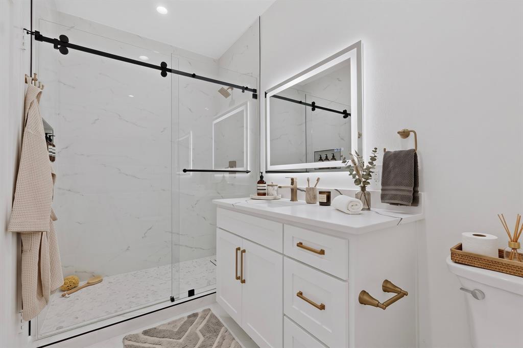 Walk-in shower with frameless glass sliding enclosure, stone-finish wall tile, white vanity featuring a stone countertop, brushed brass hardware, and a backlit wall-mounted mirror