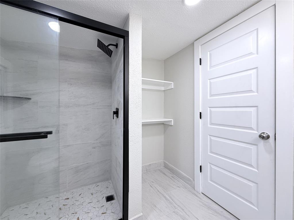 Walk-in shower featuring matte black fixtures, a rainfall showerhead, glass enclosure, and light-toned tile surfaces, accompanied by built-in wall shelving and a five-panel interior door