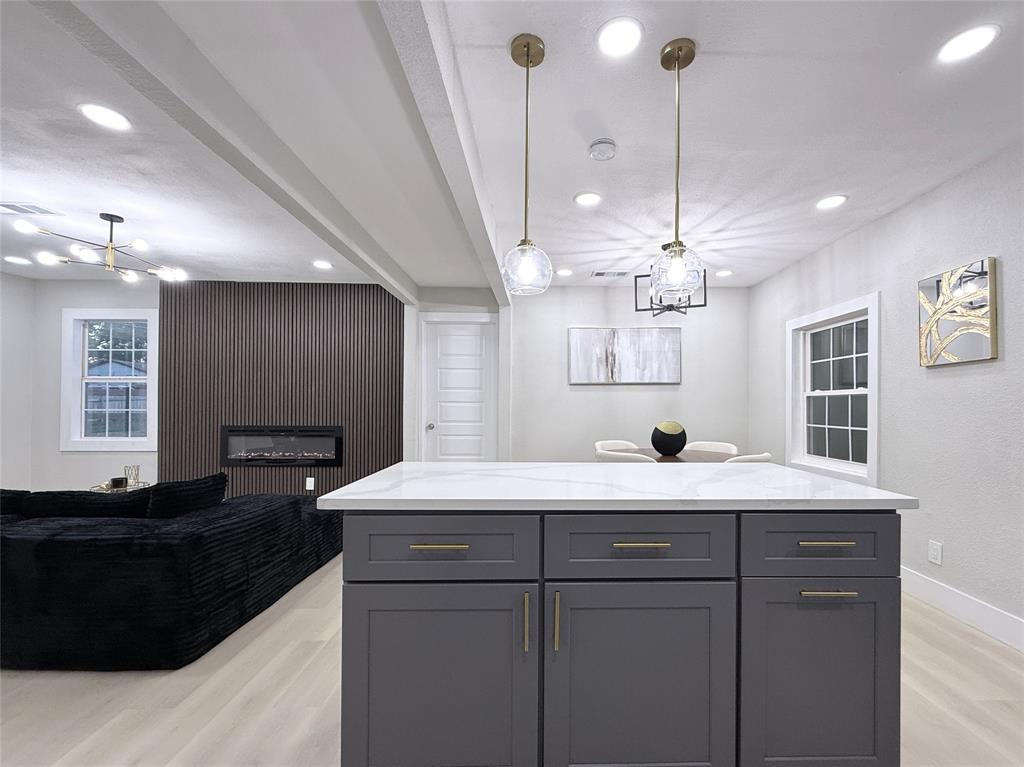 Kitchen island with quartz countertop and dark cabinetry featuring brass hardware, accented by glass pendant lighting, recessed ceiling lights, and an electric fireplace set within a vertical-slat accent wall