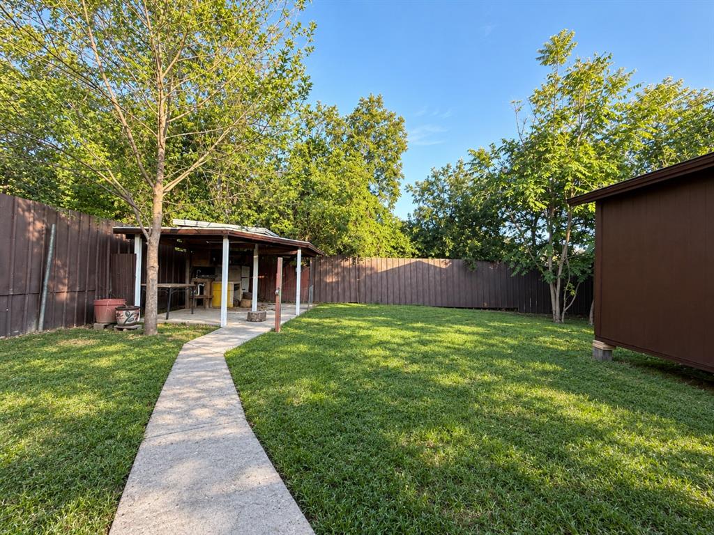 Fenced backyard featuring a manicured lawn, concrete walkway, covered patio structure, and mature trees