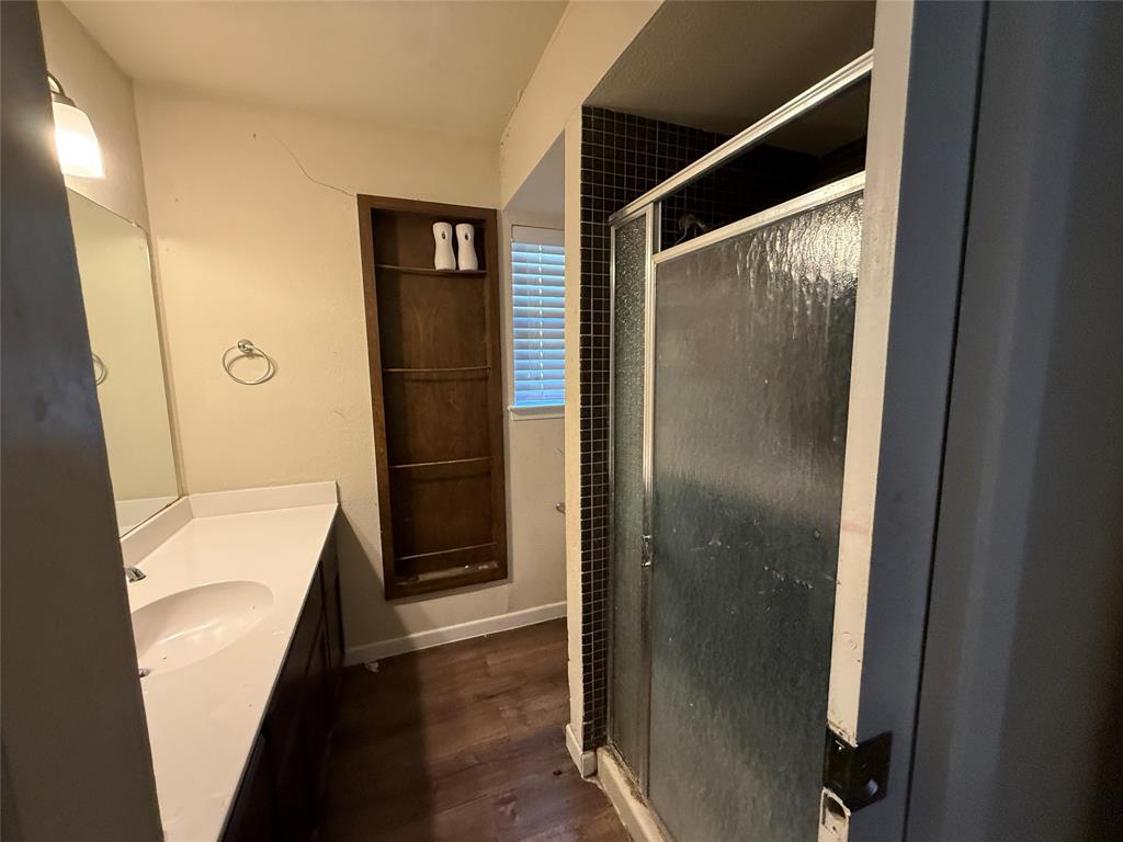 Bathroom featuring wood-finish flooring, a vanity with a white countertop and oval under-mount sink, a recessed wood-finish shelving unit, a walk-in shower with dark tile surround and textured glass door, and a wall-mounted mirror