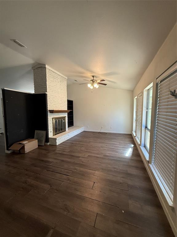 Living area featuring wood-finish flooring, vaulted ceilings, a whitewashed brick fireplace with a wood mantel, built-in shelving, and large windows with horizontal blinds
