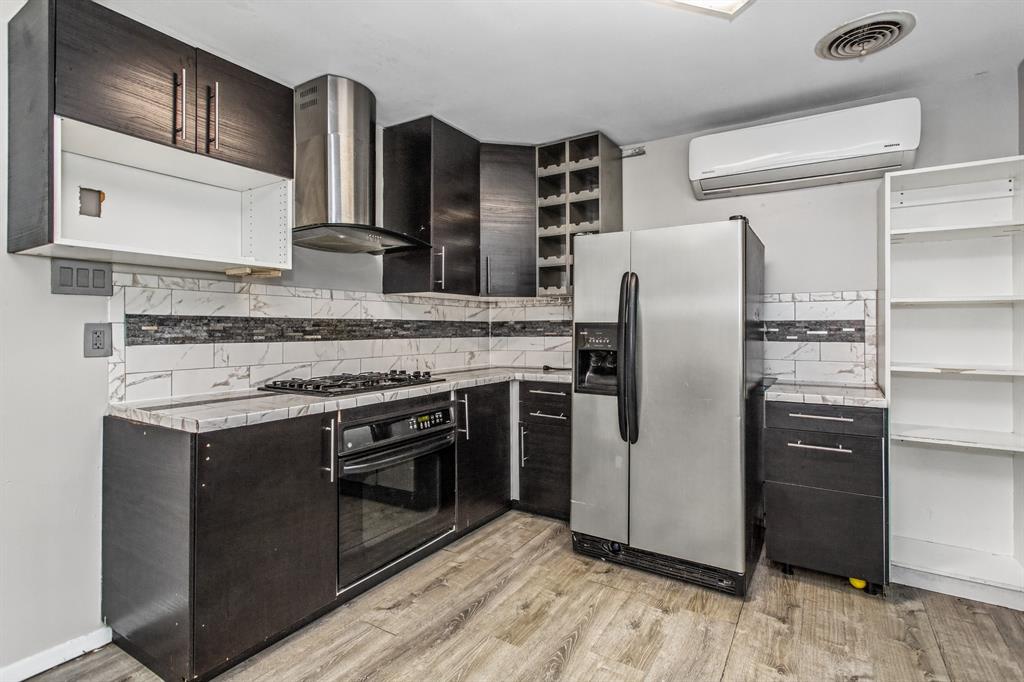 Dark-toned cabinetry featuring a built-in wine rack, stainless steel range hood, stone-patterned countertops, subway-style backsplash with decorative tile accents, and wood-finish flooring Dark-toned cabinetry featuring a built-in wine rack, stainless steel range hood, stone-patterned countertops, subway-style backsplash with decorative tile accents, and wood-finish flooring