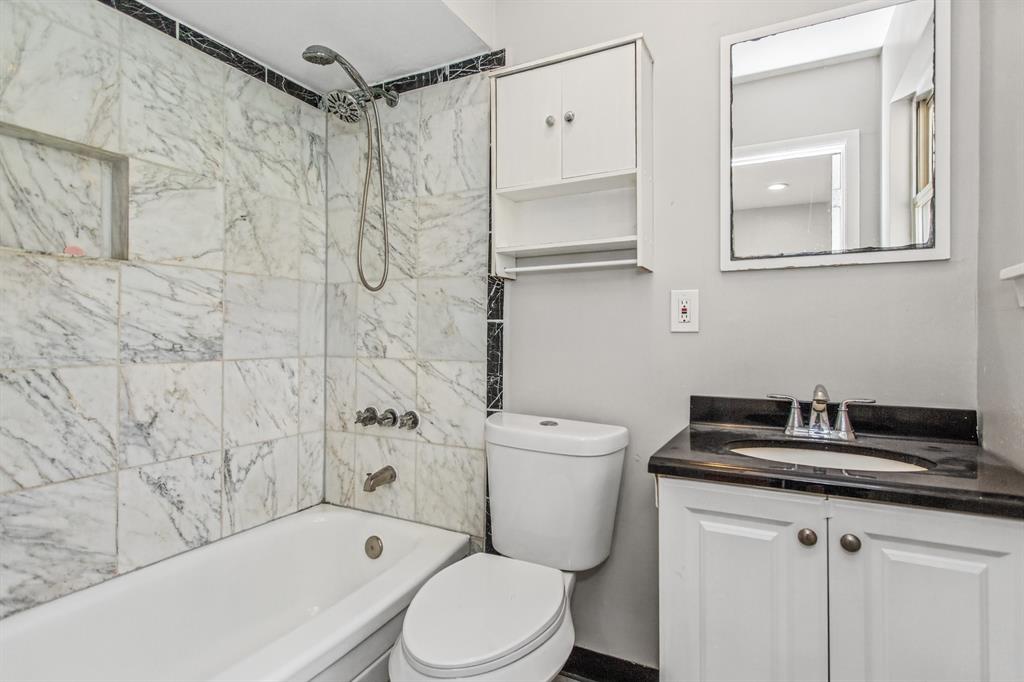 Bathroom featuring white cabinetry, dark stone vanity countertop with integrated oval sink, tub with stone-tiled surround, and wall-mounted storage cabinet