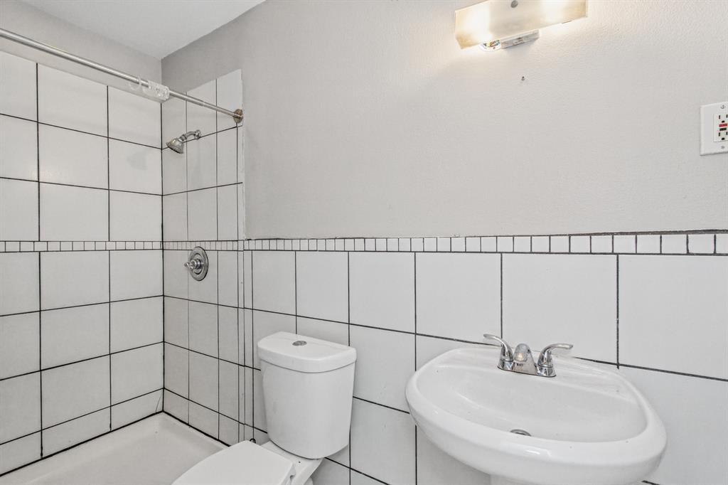 Bathroom featuring white square wall tiling with decorative mosaic accent border, pedestal sink with dual-handle chrome faucet, shower head with single-handle controls, and wall-mounted light fixture