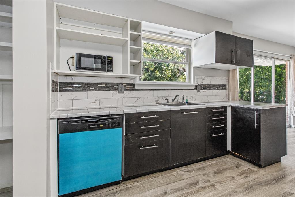 Kitchen featuring dark cabinetry, stone-pattern tile backsplash, wood-finish flooring, integrated dishwasher, and open shelving