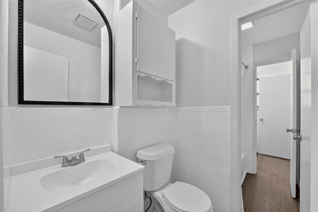White vanity with integrated sink, arched wall mirror with black trim, built-in upper cabinet, white subway tile wall surround, and wood-finish flooring