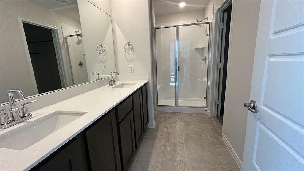 Bathroom featuring dual vanity with stone-composite countertop, dark wood cabinetry, large vanity mirror, and glass-enclosed shower with tiled flooring