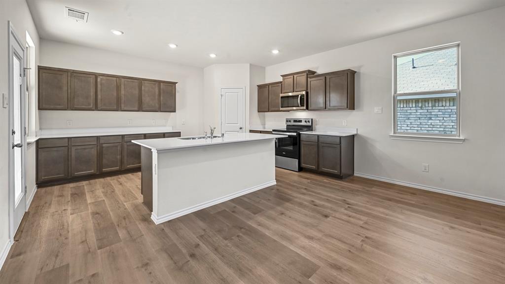Kitchen featuring wood-finish flooring, white countertops, dark wood-finish cabinetry, stainless steel appliances, and a central island with sink