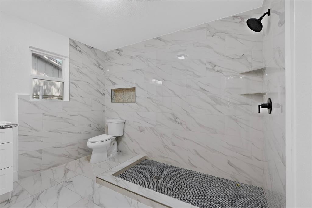 Walk-in shower featuring white large-format stone-patterned tile, matte black rainfall showerhead, integrated recessed wall niche, and dark pebble-stone flooring