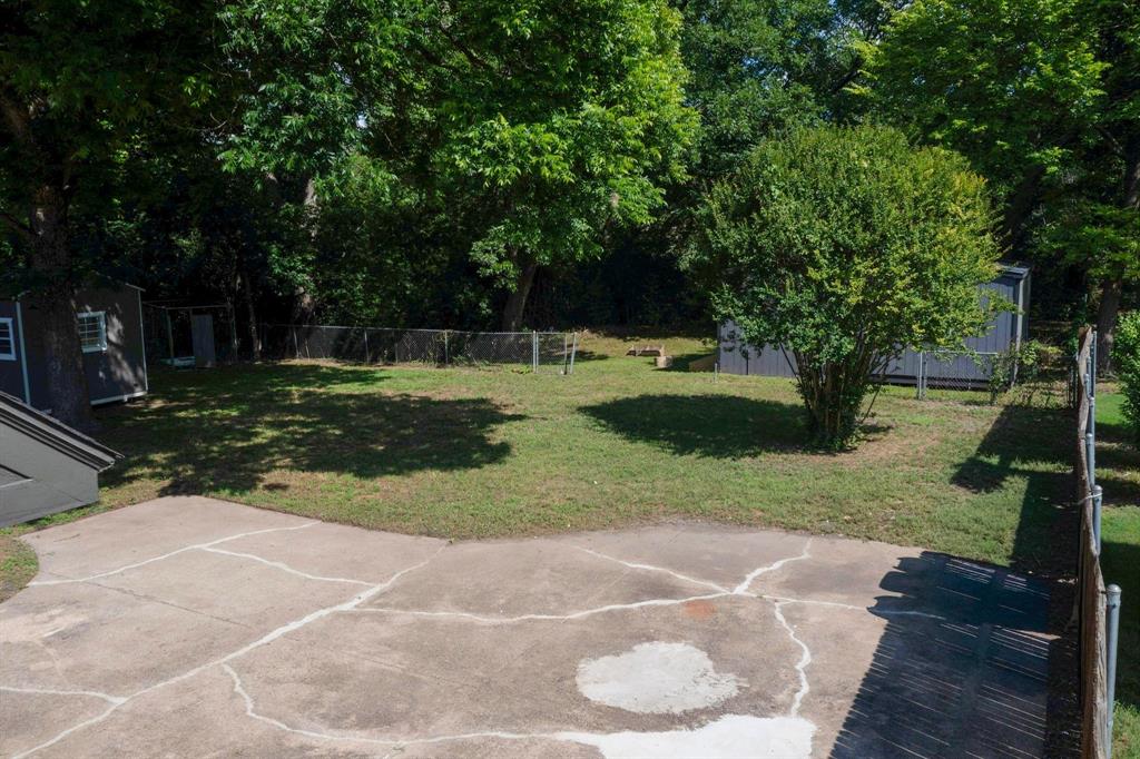 Backyard featuring a concrete patio, chain-link fencing, mature trees, and two detached storage structures