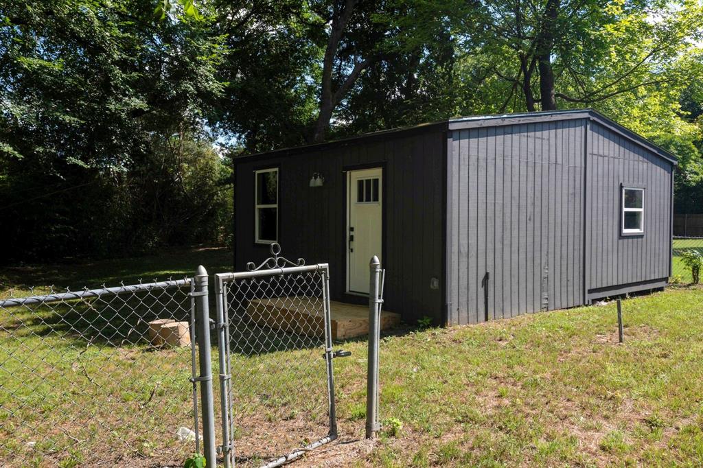 Detached structure featuring dark-toned vertical siding, a single-entry door with glass panels, multiple windows, and a surrounding chain-link fence
