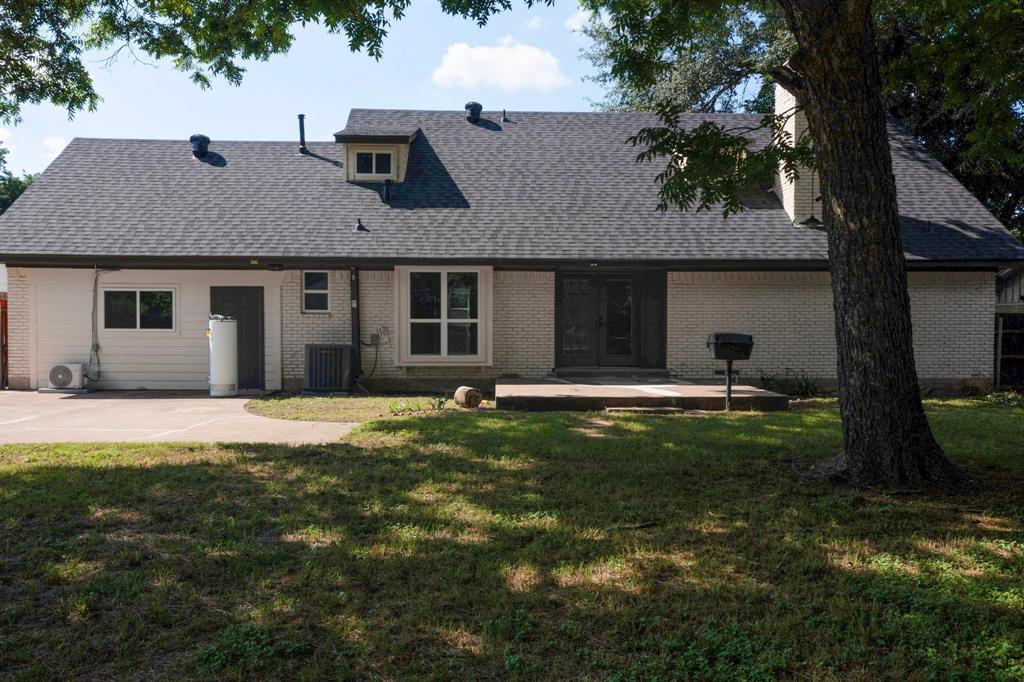 Rear exterior featuring white painted brick facade, asphalt shingle roofing, concrete patio, and mature trees