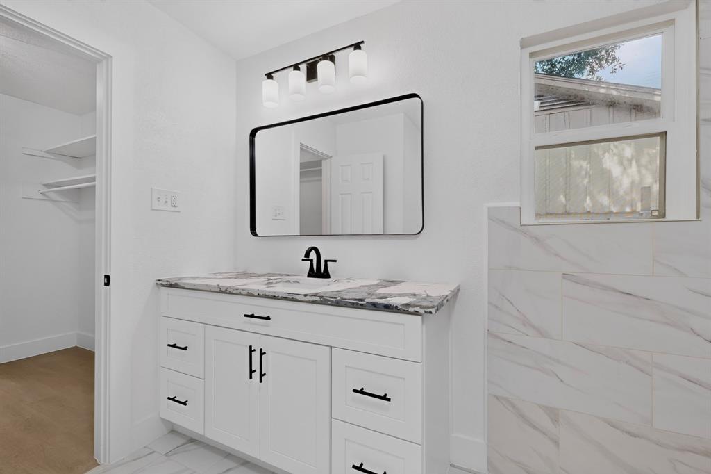 White vanity with stone-patterned countertop, rectangular undermount sink, black matte hardware, modern wall-mounted light fixture, and framed mirror