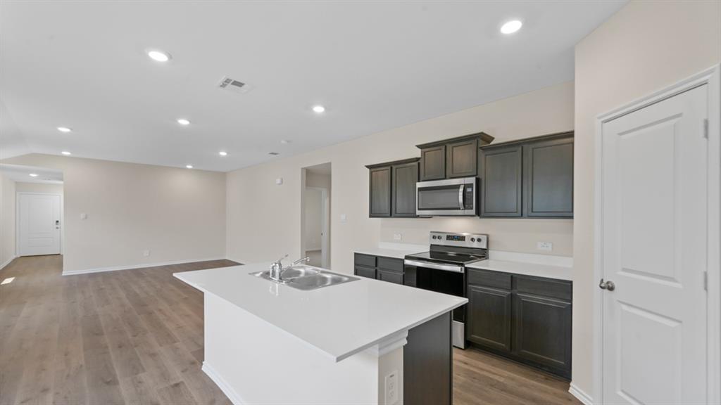 Open-concept kitchen and living area featuring wood-finish flooring, recessed lighting, a kitchen island with an integrated sink, dark wood cabinetry, and stainless steel appliances
