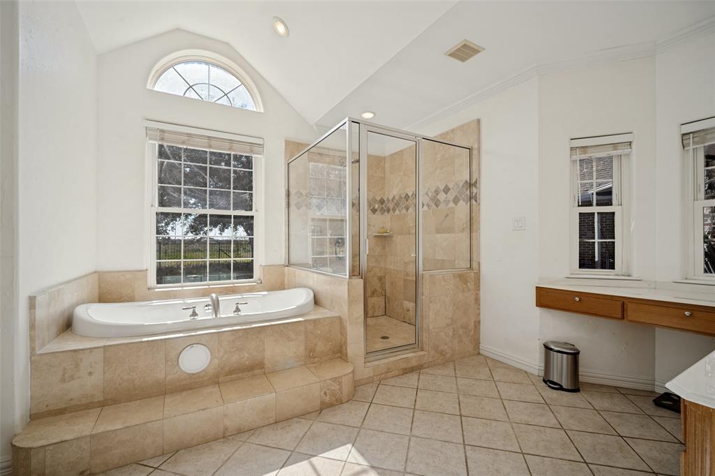 Soaking tub with stone-tile surround, glass-enclosed shower, vaulted ceiling, arched window, and dual built-in vanities.