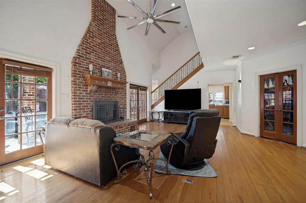 Vaulted ceiling featuring a multi-blade ceiling fan, exposed brick fireplace with a wood mantel, wood-finish flooring, staircase with wood railing, and French doors